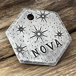 May include: A silver hexagonal pet tag with a starburst design and the word "NOVA" engraved on it.