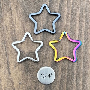 May include: Three metal star-shaped keychains in different finishes: black, silver, and rainbow. A silver disc with the text "3/4" is below the keychains.