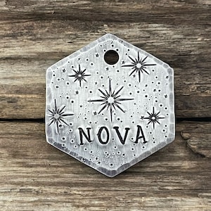 May include: A silver hexagonal pet tag with a starburst design and the word "NOVA" engraved on it. The tag has a small hole for attaching a ring.