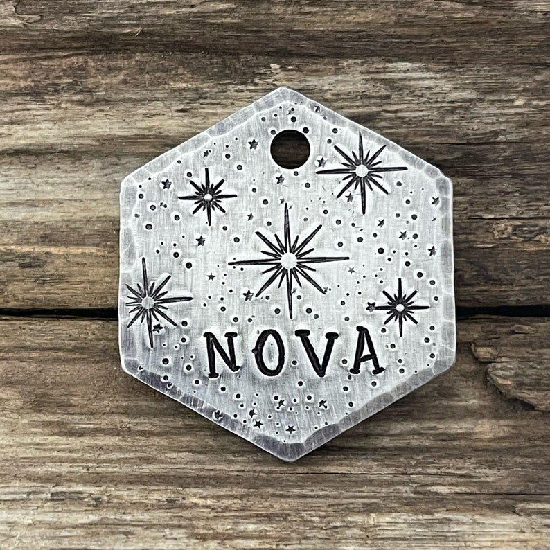 May include: A silver hexagonal pet tag with a starburst design and the word "NOVA" engraved on it. The tag has a small hole for attaching a ring.
