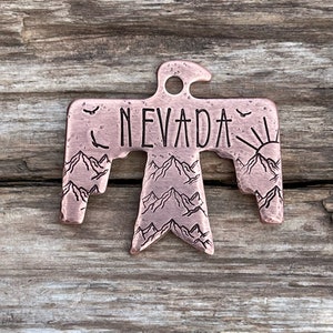 Puede incluir: Adorno de metal de color cobre con forma de pájaro estilizado con la palabra "NEVADA" inscrita en el pecho. El pájaro tiene un diseño de cordillera en sus alas y un sol saliendo sobre las montañas en el ala derecha.