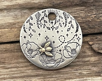 Puede incluir: Un amuleto de plata con una abeja volando sobre un diseño floral. La abeja está hecha de metal dorado y las flores están delineadas en negro. El amuleto tiene un agujero en la parte superior para sujetarlo a un collar o pulsera.