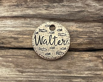 May include: A round, brass dog tag with a hammered finish. The tag is decorated with a pattern of small fish and the name "Walter" is engraved in a cursive font.