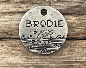 May include: A silver round pet tag engraved with the name "BRODIE". The tag features a stylised image of a fish jumping out of the water with waves and raindrops.