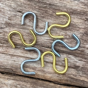 Puede incluir: Un surtido de ganchos en forma de S en tonos dorados y plateados. Los ganchos metálicos están dispuestos sobre una superficie de madera envejecida. Estos ganchos están diseñados para colgar objetos y son un accesorio de ferretería versátil.