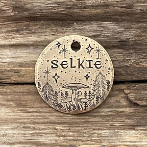 May include: A round brass tag with an engraved design of a forest scene with mushrooms, stars, and the word "Selkie".
