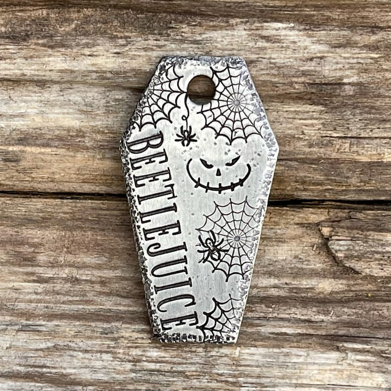 Puede incluir: Un amuleto de metal plateado en forma de ata&uacute;d con un dise&ntilde;o de telara&ntilde;a y las palabras "Beetlejuice" grabadas en el lateral. El amuleto tiene un peque&ntilde;o agujero en la parte superior para sujetarlo a un collar o pulsera.