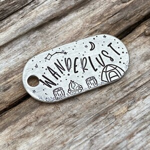 May include: A silver oval-shaped keychain with the word "wanderlust" engraved on it. The keychain also features a small tent, campfire, and two trees.