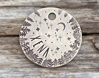 May include: A round brass tag with a hole for a ring. The tag has a fern design around the edge and a crescent moon and stars in the center. The name "Yena" is engraved in the center of the tag.
