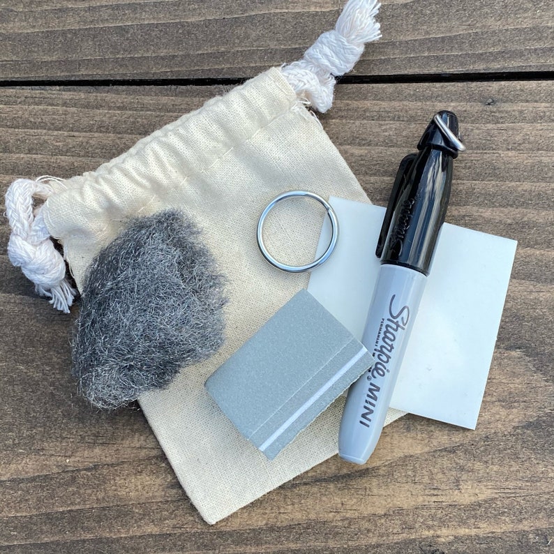 May include: A small white canvas bag with a grey scrubby, a silver key ring, a grey rectangular sanding block, and a black Sharpie Mini permanent marker.