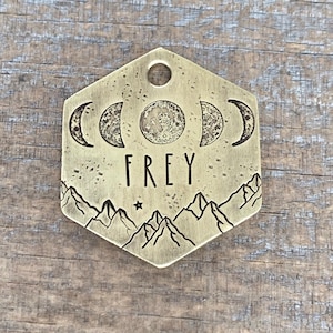 May include: Brass hexagonal dog tag with a moon phase design, mountains, and the name "FREY" engraved on it.