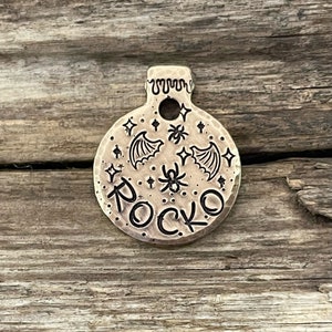 May include: A round, brass dog tag with a black engraved design of bats, spiders, and stars. The name "ROCKO" is engraved in a gothic font.