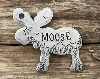 May include: Silver moose-shaped pet tag with a mountain and forest scene engraved inside the moose silhouette. The name "MOOSE" is engraved on the tag.