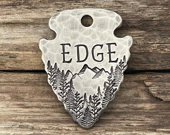May include: Silver arrowhead-shaped metal tag with the word "EDGE" engraved on it. The tag features a mountain scene with trees engraved below the word.