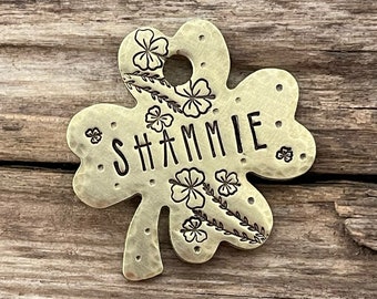 May include: A gold-coloured metal shamrock-shaped pet tag with the name "SHAMMIE" engraved in black lettering. The tag is decorated with small, engraved shamrocks and vines.