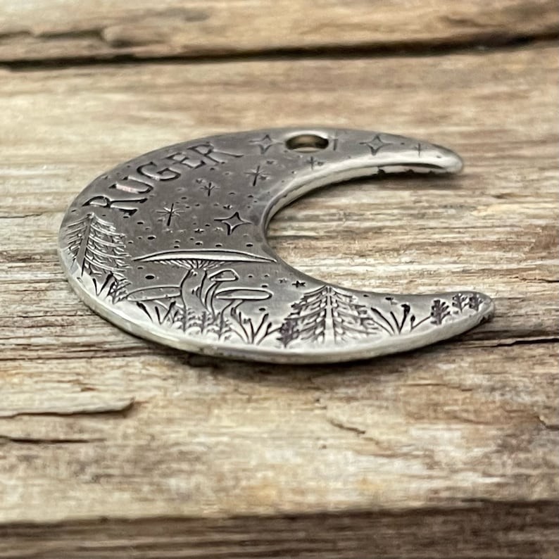 May include: A silver crescent moon charm with the name "RUGER" engraved on it. The charm features a detailed design of a forest scene with trees, mushrooms, and stars.