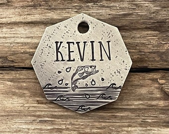 May include: A silver-toned, octagonal dog tag with a black engraved design of a fish jumping out of the water with waves and raindrops. The name "KEVIN" is engraved above the fish.