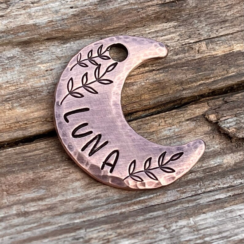 Op de afbeelding: Een koperkleurige, halve maanvormige huisdiertag met de naam "LUNA" erop gegraveerd. De tag is aan beide kanten van de naam versierd met een bladrankenontwerp.