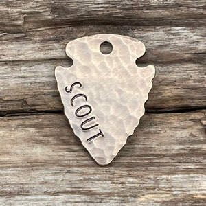 Op de afbeelding: Een zilveren hondenpenning in de vorm van een pijlpunt met het woord "SCOUT" erop gegraveerd.