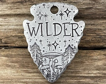 May include: A silver-colored, arrowhead-shaped tag with the word "WILDER" engraved in bold letters. The tag features a forest scene with trees, mushrooms, and stars. A small hole is at the top for attachment.