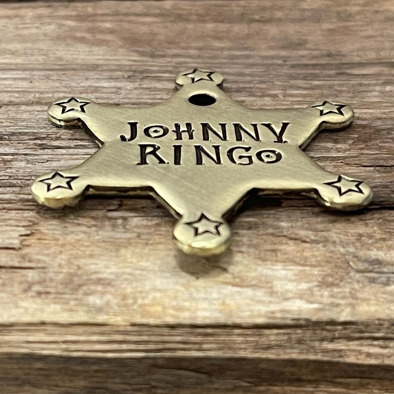 Peut inclure: Une &eacute;toile de sh&eacute;rif dor&eacute;e &agrave; cinq branches avec un trou au centre. L'&eacute;toile porte la gravure "JOHNNY RINGO".