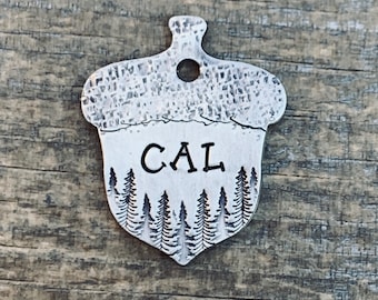 May include: Silver acorn-shaped pet tag featuring a forest scene and the name "CAL" engraved on it.