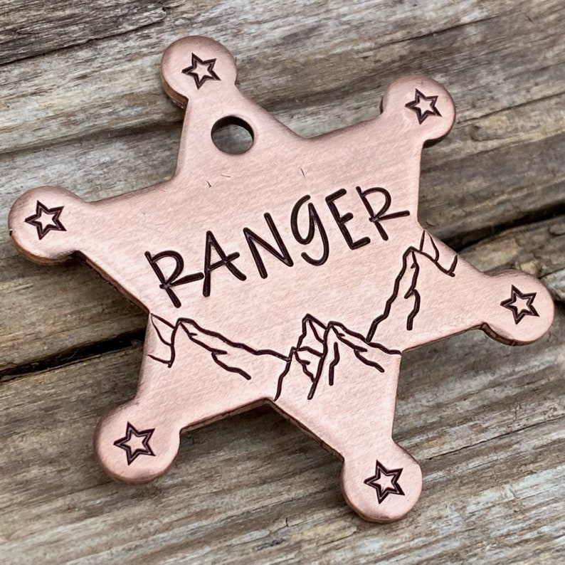 May include: A copper-colored sheriff's star-shaped dog tag with the word "RANGER" engraved on it. The tag also features a mountain range design.