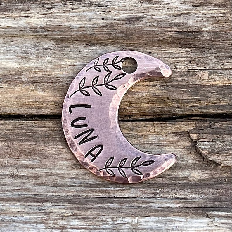 Op de afbeelding: Een koperkleurige halve maan hondenhanger met de naam "LUNA" erop gegraveerd. De hanger heeft een gehamerde textuur en is versierd met kleine bladeren.