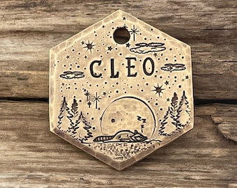 Può includere: Un'etichetta esagonale in ottone per animali domestici con un design di un procione che dorme, una luna piena, stelle e nuvole. L'etichetta è incisa con il nome "CLEO".