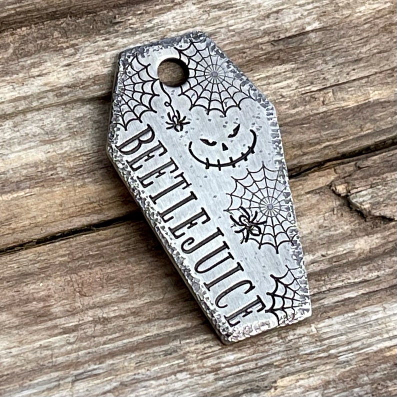 Puede incluir: Un llavero de metal plateado en forma de ata&uacute;d con las palabras "Beetlejuice" grabadas en &eacute;l. El llavero est&aacute; decorado con un dise&ntilde;o de telara&ntilde;a y una cara de calabaza espeluznante.