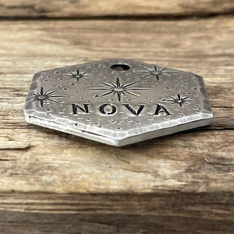 May include: A silver hexagonal charm with a starburst design and the word "NOVA" engraved on it.