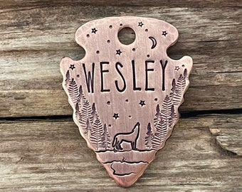 May include: A copper-colored arrowhead-shaped tag with the name "WESLEY" engraved in the center. The tag features a wolf howling at the moon, surrounded by trees and stars.