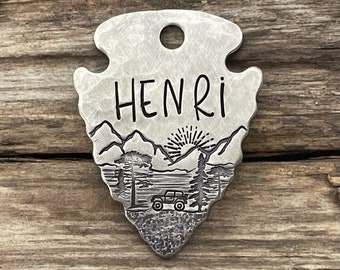May include: Silver arrowhead-shaped tag with the name "HENRI" engraved on it. The tag features a mountain scene with a jeep driving through it and a sun rising over the mountains.