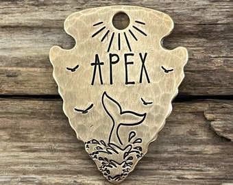 May include: A brass arrowhead-shaped keychain with a whale tail design, the word "APEX" and a sun with rays.