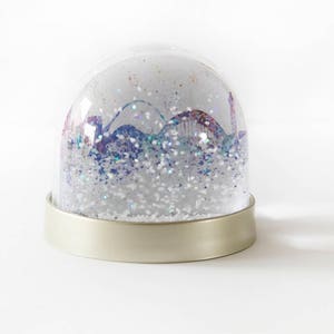 Op de afbeelding: Een transparante glazen sneeuwbol met een zilveren basis. De bol bevat glitter en een kleurrijk stadsbeeld van binnen.