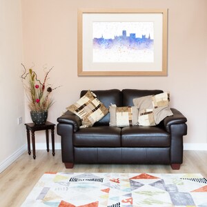 Peut inclure: Un canap&eacute; en cuir marron avec deux coussins, un tableau encadr&eacute;e d'une aquarelle repr&eacute;sentant un horizon urbain, et une petite table avec une plante en pot dans un salon avec une chemin&eacute;e et un tapis.