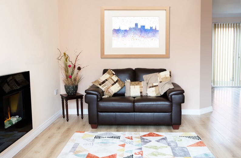 Peut inclure: Un canap&eacute; en cuir marron avec deux coussins, un tableau encadr&eacute;e d'une aquarelle repr&eacute;sentant un horizon urbain, et une petite table avec une plante en pot dans un salon avec une chemin&eacute;e et un tapis.