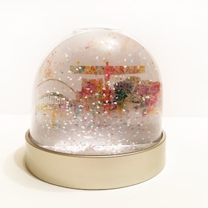 Puede incluir: Un globo de nieve de vidrio con una base dorada. El globo contiene una imagen colorida de un horizonte de ciudad con un puente y una gran estructura en el fondo. El globo de nieve está lleno de purpurina blanca.