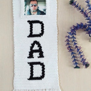 May include: White crocheted wall hanging with black letters spelling "DAD" and a framed photo in the top center. The hanging has white fringe at the bottom.