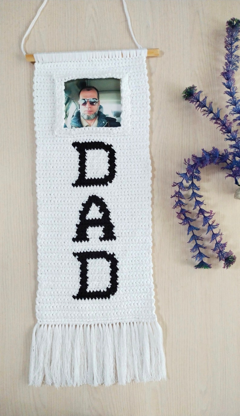 May include: White crocheted wall hanging with black letters spelling "DAD" and a framed photo in the top center. The hanging has white fringe at the bottom.