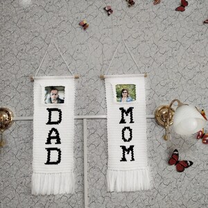 May include: Two white crocheted wall hangings with black lettering spelling "DAD" and "MOM". Each hanging has a small framed photo of a person and a fringe at the bottom.