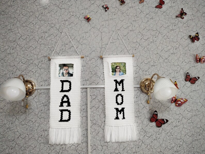 May include: Two white crocheted wall hangings with black lettering spelling "DAD" and "MOM". Each hanging has a small framed photo of a person and a fringe at the bottom.