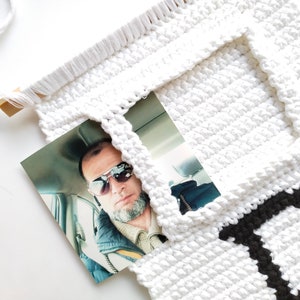 May include: White crocheted wall hanging with a black letter "D" and a photo of a man wearing sunglasses in a car.