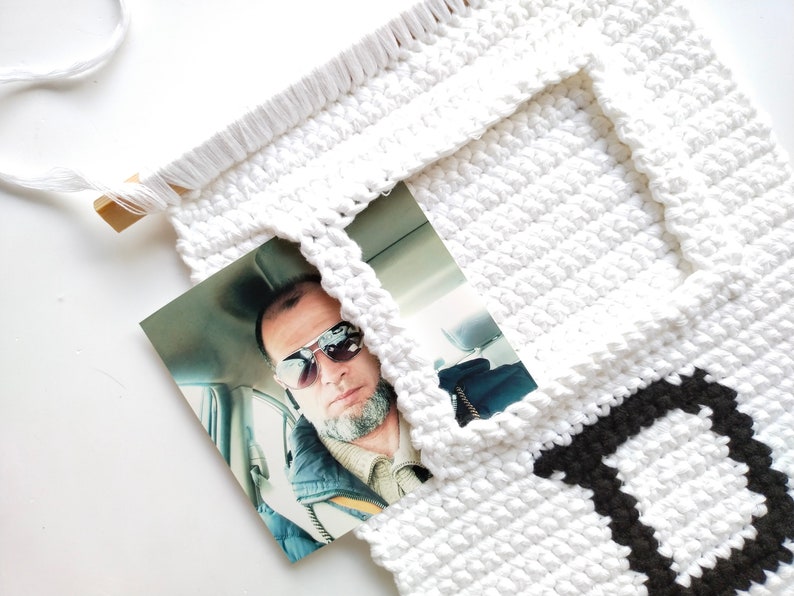 May include: White crocheted wall hanging with a black letter "D" and a photo of a man wearing sunglasses in a car.