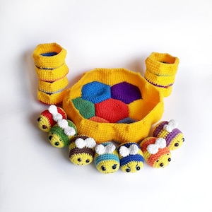 Op de afbeelding: Een gehaakt bijenkorfspel met zes kleurrijke bijen en twee gele manden. De bijenkorf is gemaakt van geel garen met een veelkleurig honingraatcentrum. De bijen zijn geel met zwarte strepen en witte vleugels.
