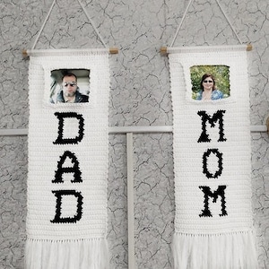May include: Two white crocheted wall hangings with black lettering. The left hanging says "DAD" and the right hanging says "MOM". Each hanging has a small photo frame with a picture of a person inside.