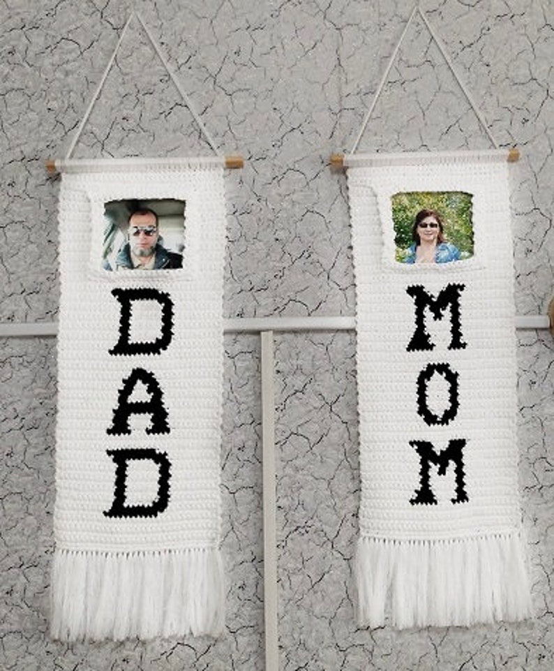 May include: Two white crocheted wall hangings with black lettering. The left hanging says "DAD" and the right hanging says "MOM". Each hanging has a small photo frame with a picture of a person inside.