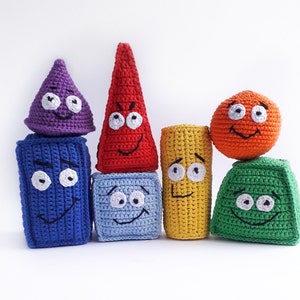 Puede incluir: Un conjunto de seis juguetes de peluche tejidos a crochet con forma de figuras geométricas y caras sonrientes. Los juguetes son de los colores del arcoíris: morado, rojo, amarillo, verde, azul y naranja.