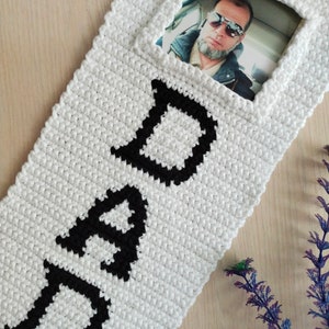 May include: White crocheted wall hanging with the word "DAD" in black yarn. A photo of a man wearing sunglasses is in a frame on the hanging.