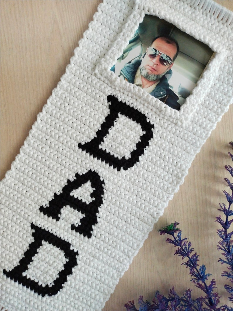 May include: White crocheted wall hanging with the word "DAD" in black yarn. A photo of a man wearing sunglasses is in a frame on the hanging.